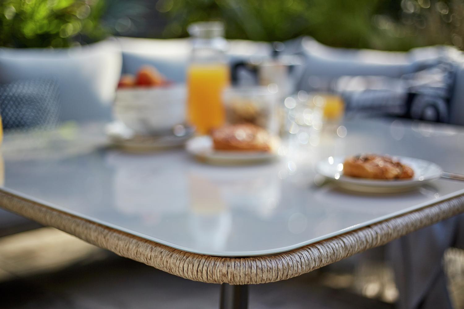 Outdoor dining table with glass top and woven edge, set for breakfast with pastries and juice