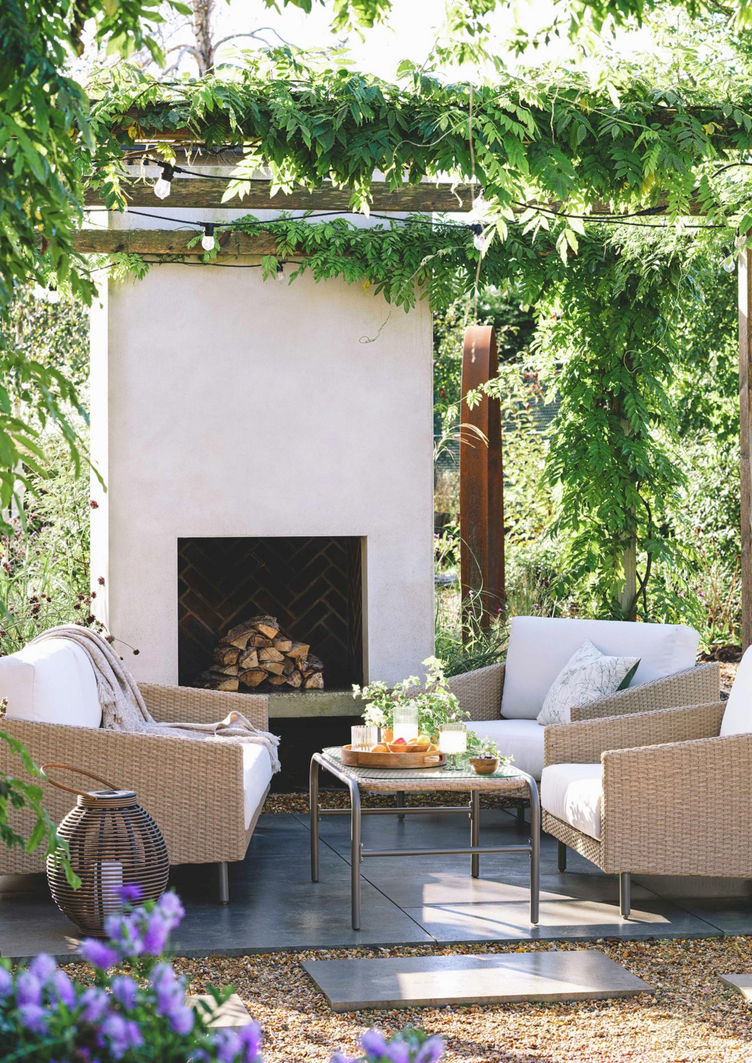 Luxury outdoor wicker sofa set with white cushions, glass table, and fireplace under leafy pergola