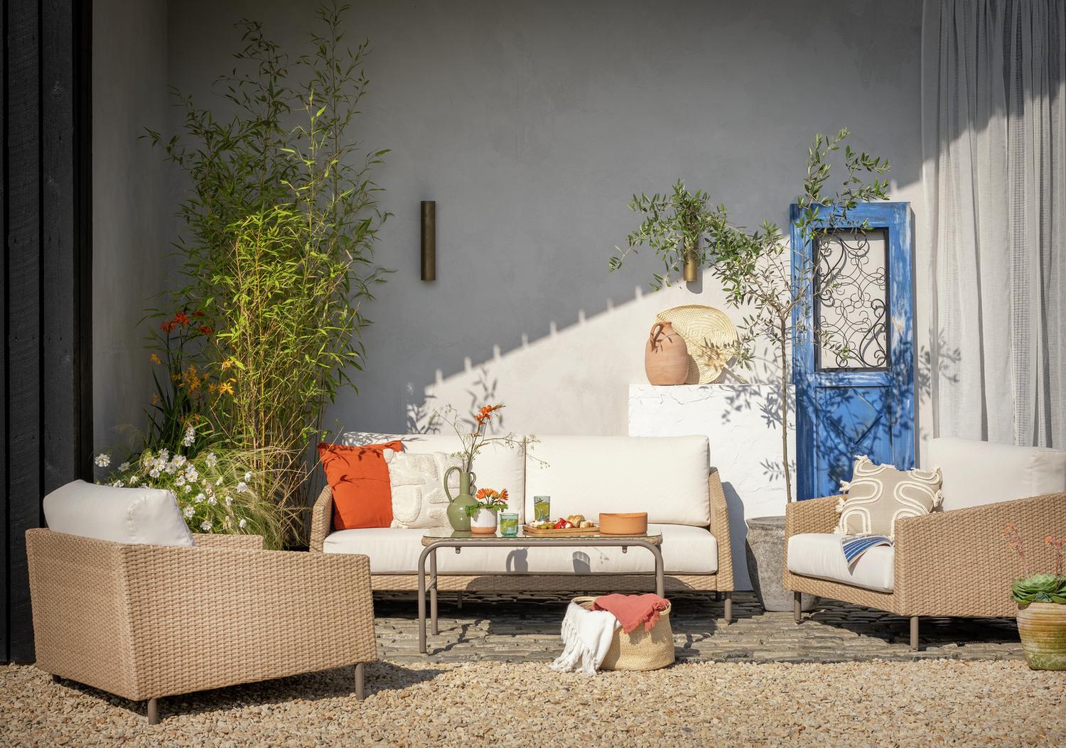Luxury outdoor wicker sofa set with white cushions, orange throw pillows, and glass coffee table on gravel patio