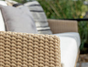 Woven outdoor sofa with beige rattan frame, white cushions, and grey throw pillows in garden setting