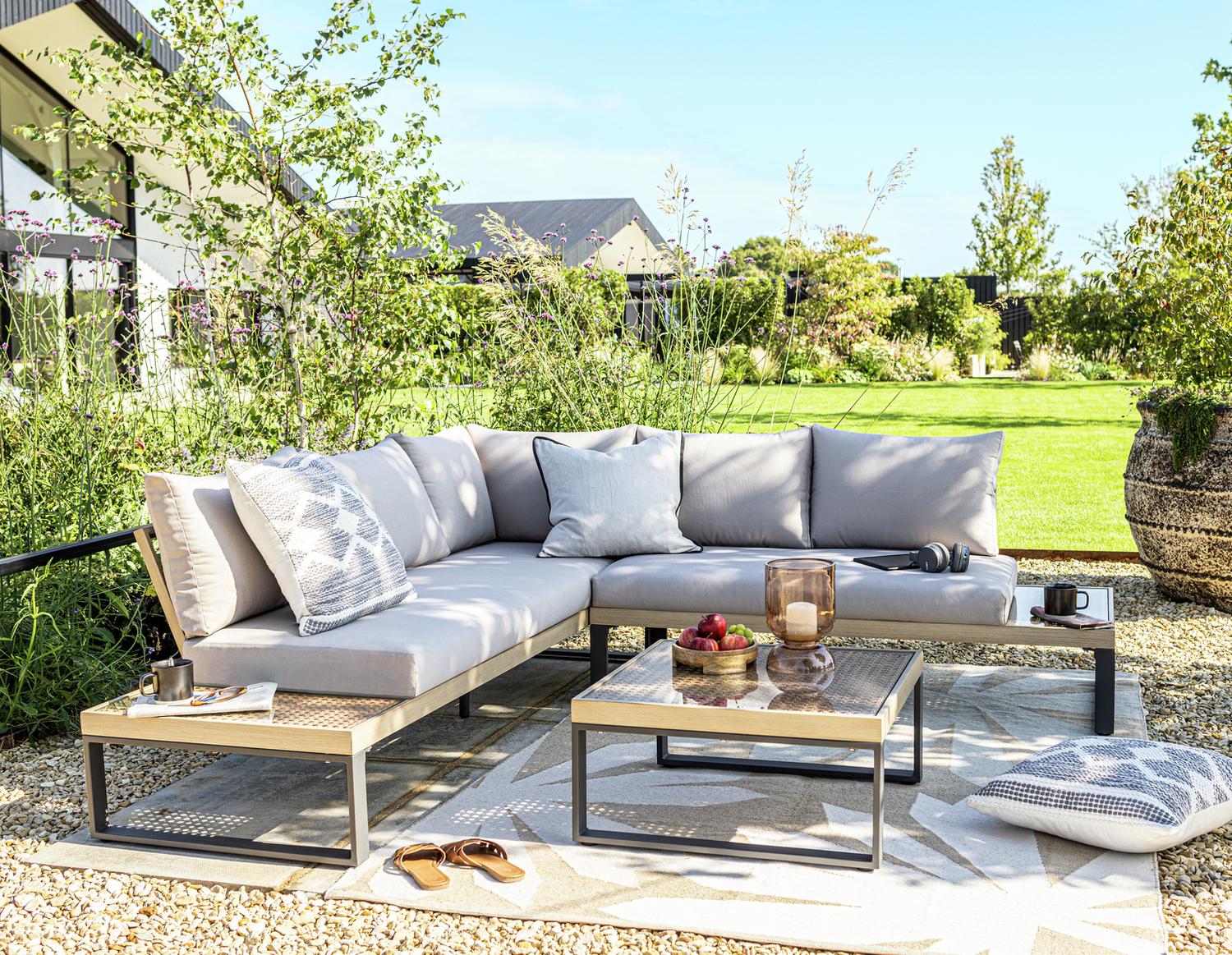 Luxury outdoor sectional sofa with grey cushions, glass coffee table, and patterned rug in a sunny garden setting