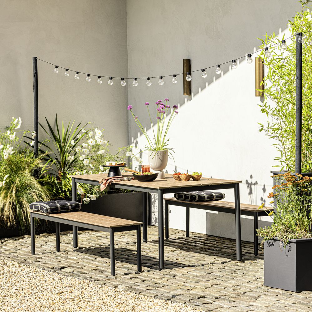 Modern outdoor dining set with wooden table, benches, string lights, and potted plants on stone patio