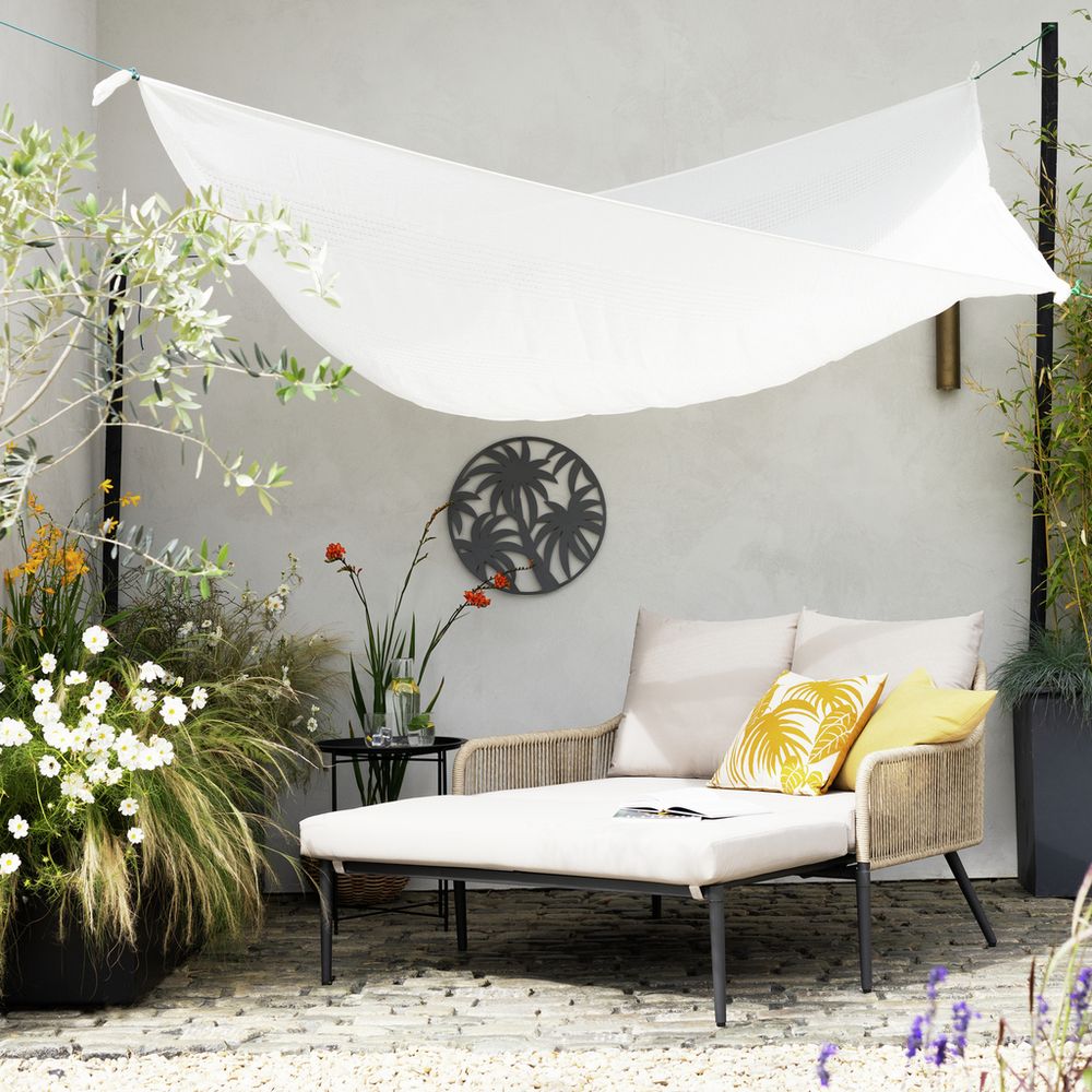 Outdoor wicker daybed with white cushions under shade sail, surrounded by garden plants and wall art