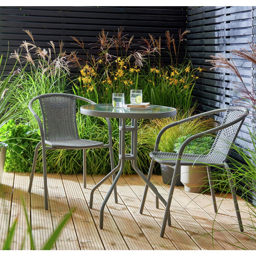 Grey wicker bistro set with glass table on wooden deck, surrounded by greenery and flowers