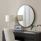 Round black-framed wall mirror above dark wood vanity with lamp, photo frame, and plant in modern bedroom