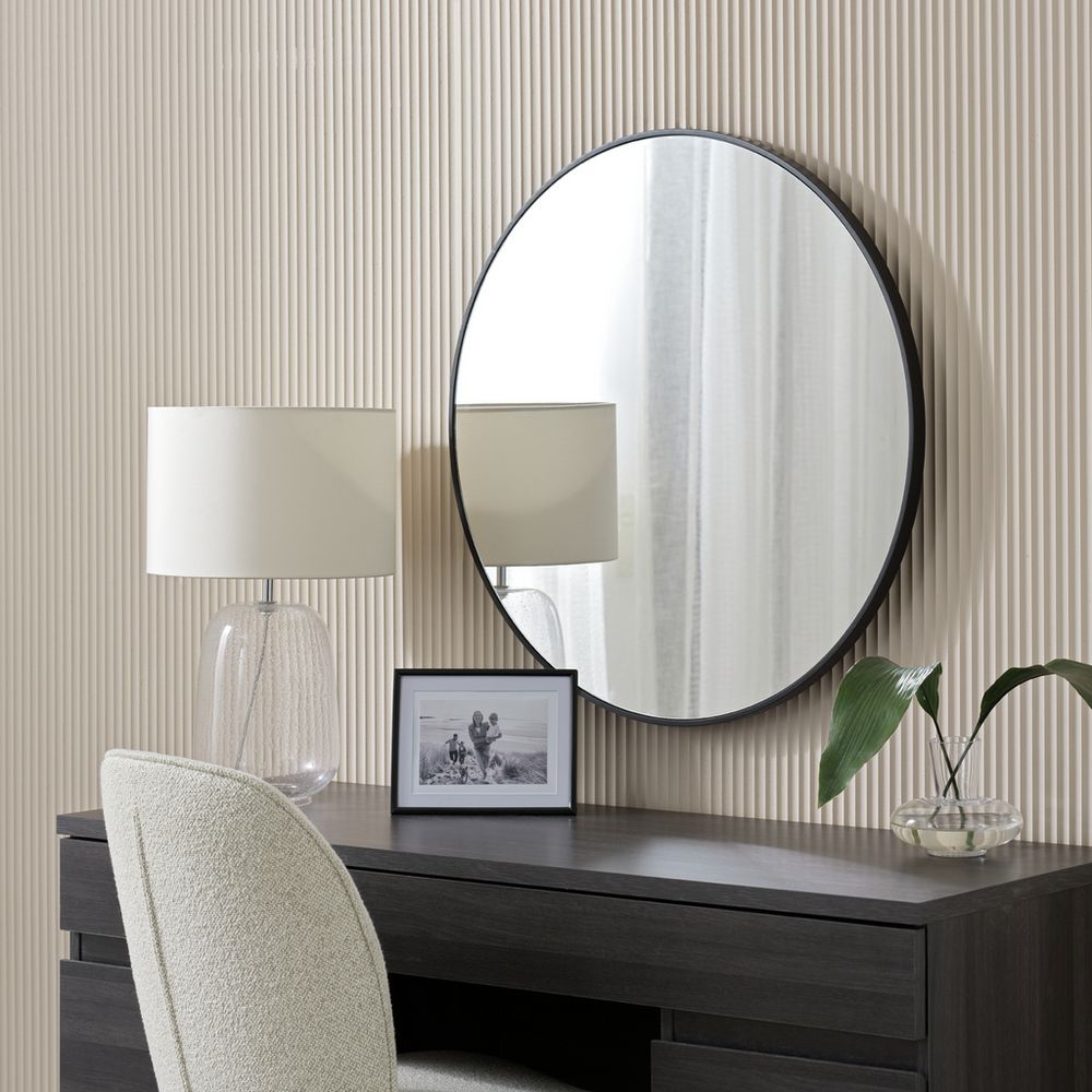 Round black-framed wall mirror above dark wood vanity with lamp, photo frame, and plant in modern bedroom