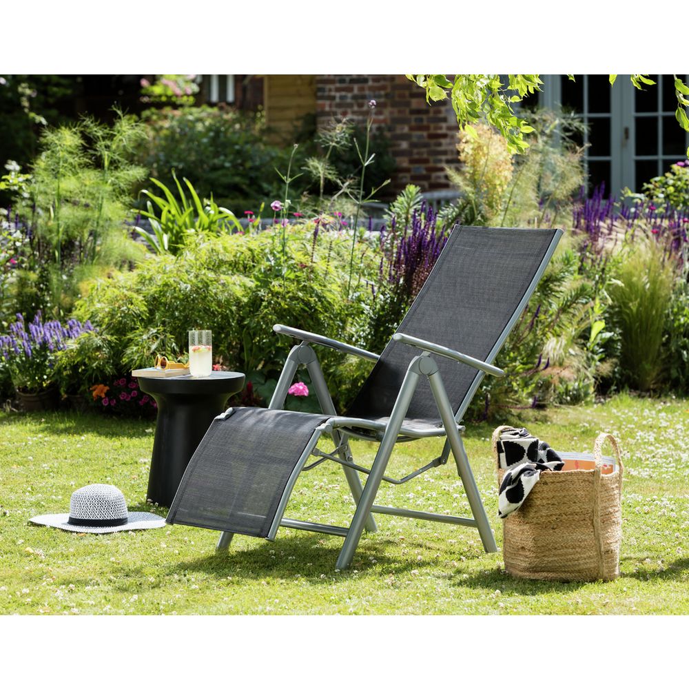 Black outdoor recliner chair on grass with side table, hat, and basket in garden setting