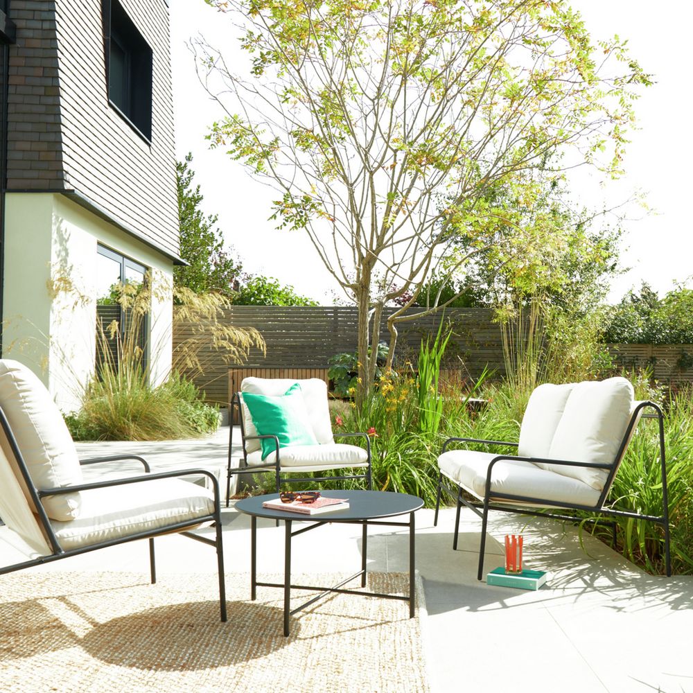 Modern outdoor patio furniture set with white cushions, black metal frames, and a round coffee table in a sunny garden setting.