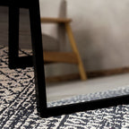 Black-framed mirror on patterned rug, reflecting wooden chair in modern living room