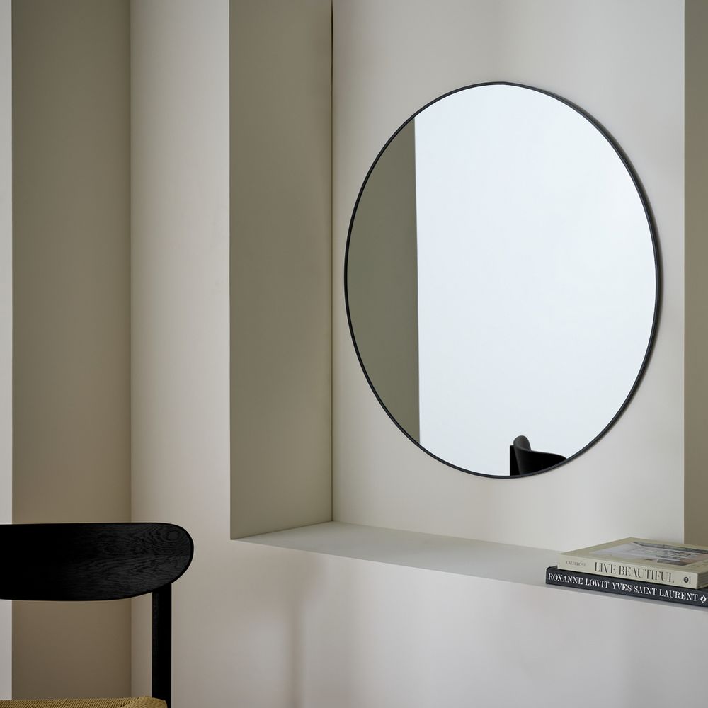 Modern round wall mirror with black frame in minimalist living space, styled with books and chair