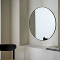 Modern round wall mirror with black frame in minimalist living space, styled with books and chair
