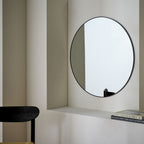 Modern round wall mirror with black frame in minimalist living space, styled with books and chair