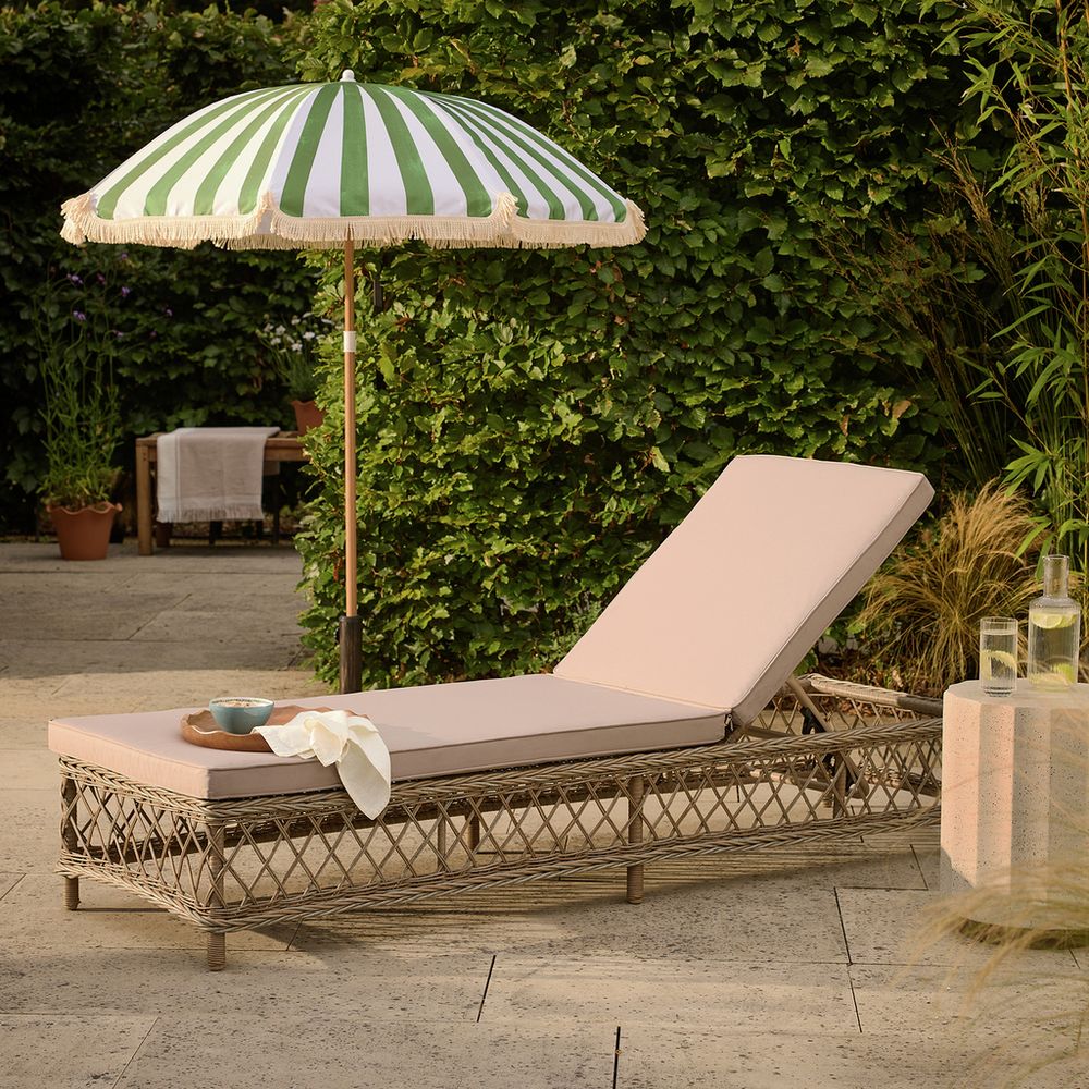 Wicker outdoor chaise lounge with beige cushion under green and white striped umbrella in garden setting