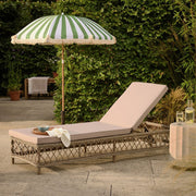 Wicker outdoor chaise lounge with beige cushion under green and white striped umbrella in garden setting