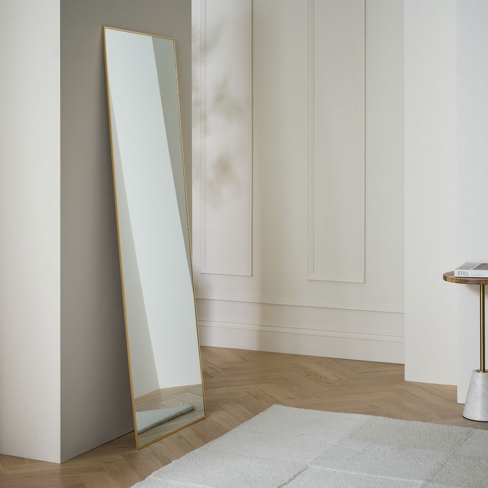 Full-length gold-framed mirror leaning against wall in modern living room with herringbone wood floor