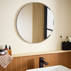 Round bathroom mirror with black frame above wooden vanity, modern sink faucet, and toiletries