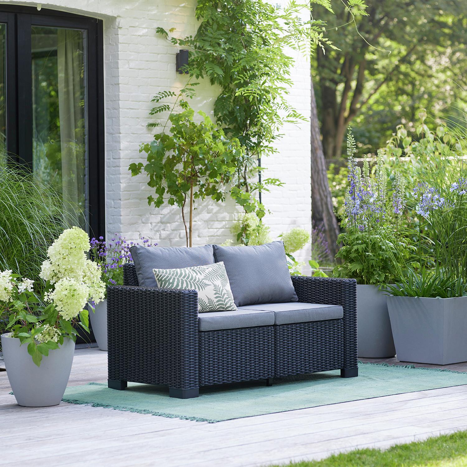Dark wicker outdoor sofa with grey cushions on patio, surrounded by greenery and potted plants
