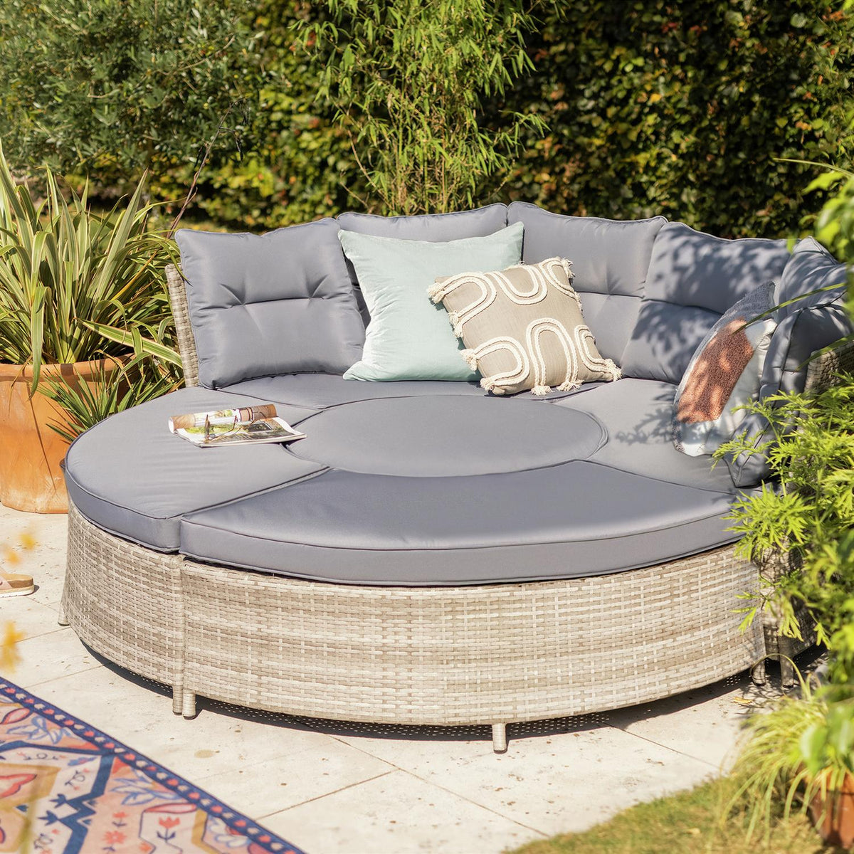 Round outdoor wicker sofa with grey cushions and decorative pillows in garden setting