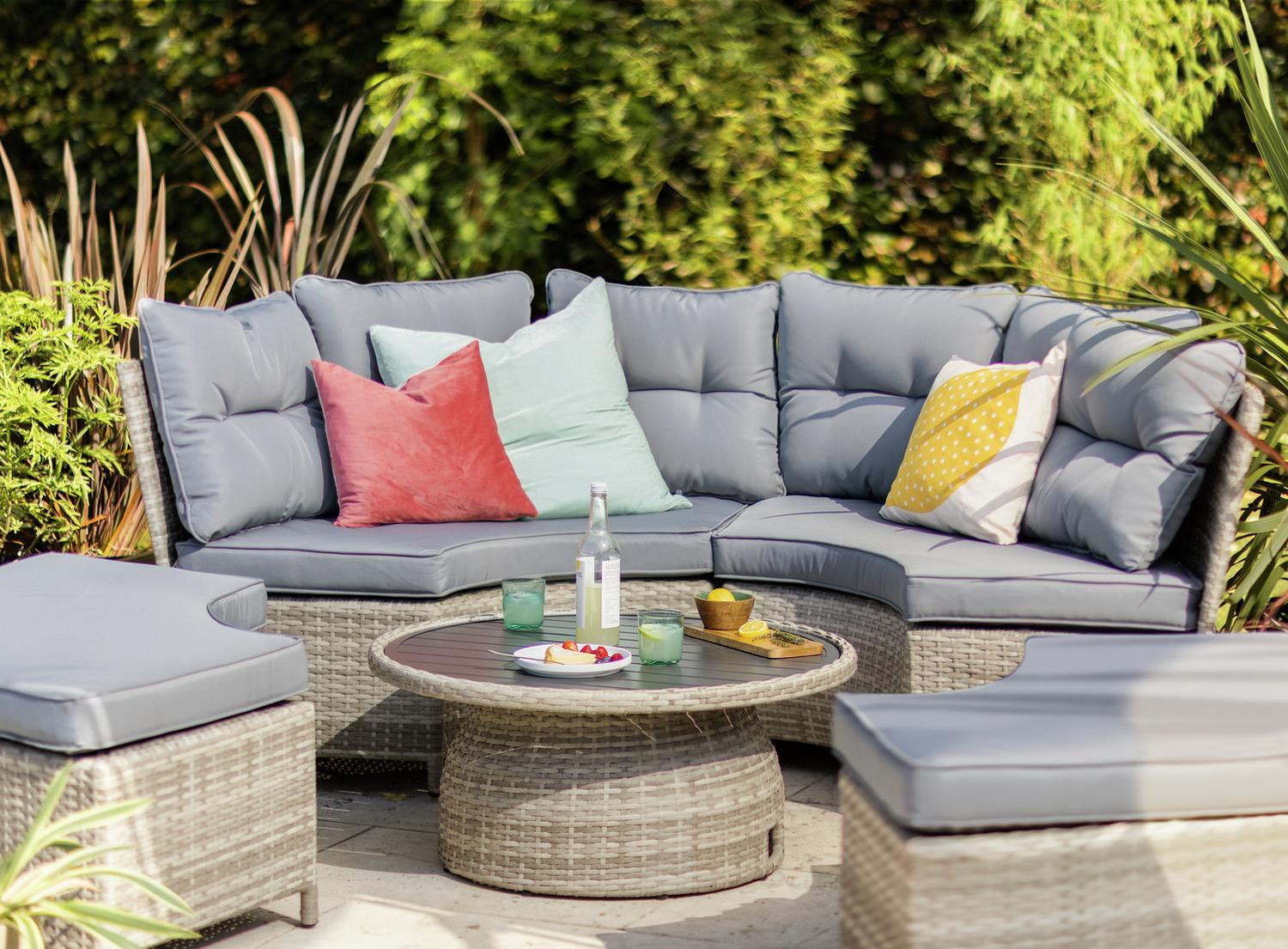 Grey wicker outdoor sofa set with colorful cushions, round table, and garden backdrop