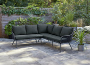 Dark green outdoor corner sofa with woven frame on patio, surrounded by plants and garden greenery