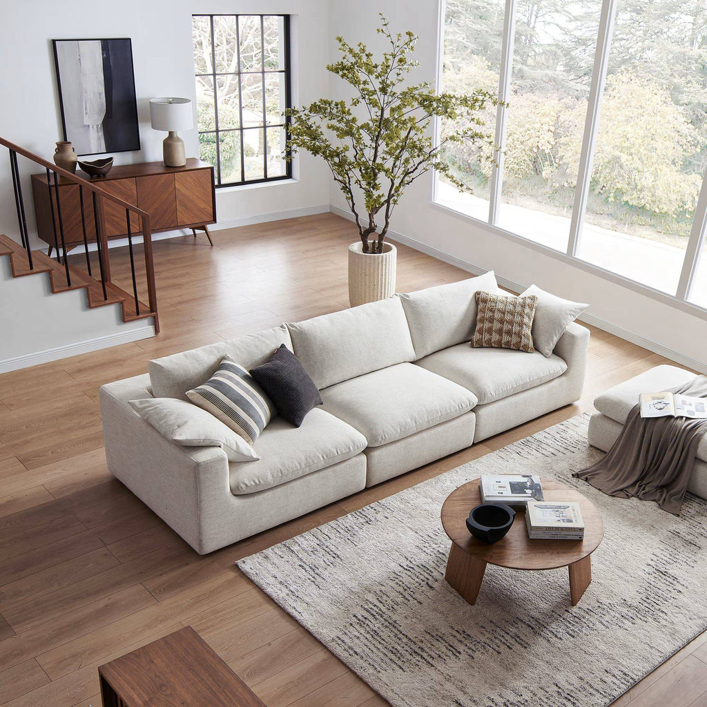 Modern living room with beige extended sofa and ottoman, large windows, and neutral decor
