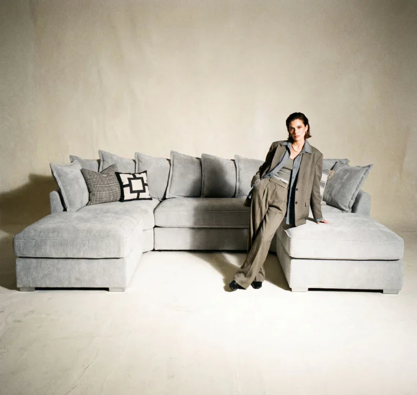 Modern grey corner sofa with scatter cushions in a minimalist living room setting, person posing