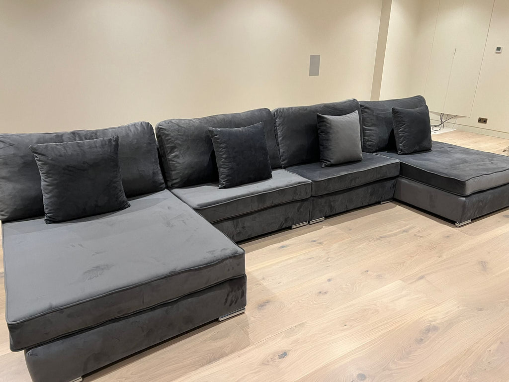 Modern grey velvet corner sofa with five cushions on light wood flooring in a minimalist room