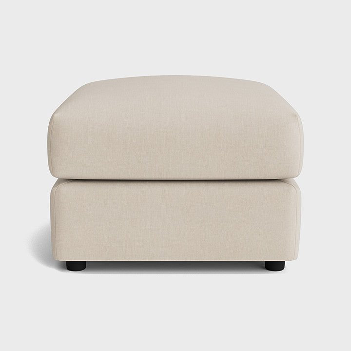 Cream fabric footstool with soft cushioning and black feet on a white background