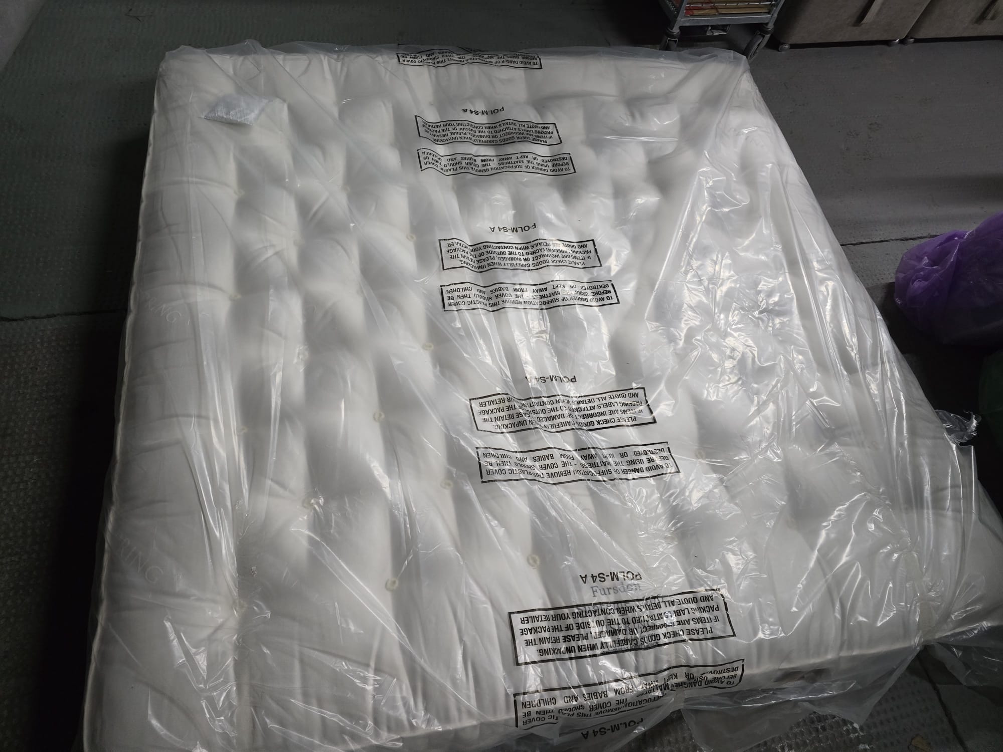 Emperor size white mattress in protective plastic wrap on a grey floor