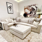 Modern oatmeal corner sofa with ottoman in a stylish living room, featuring neutral decor and abstract wall art.