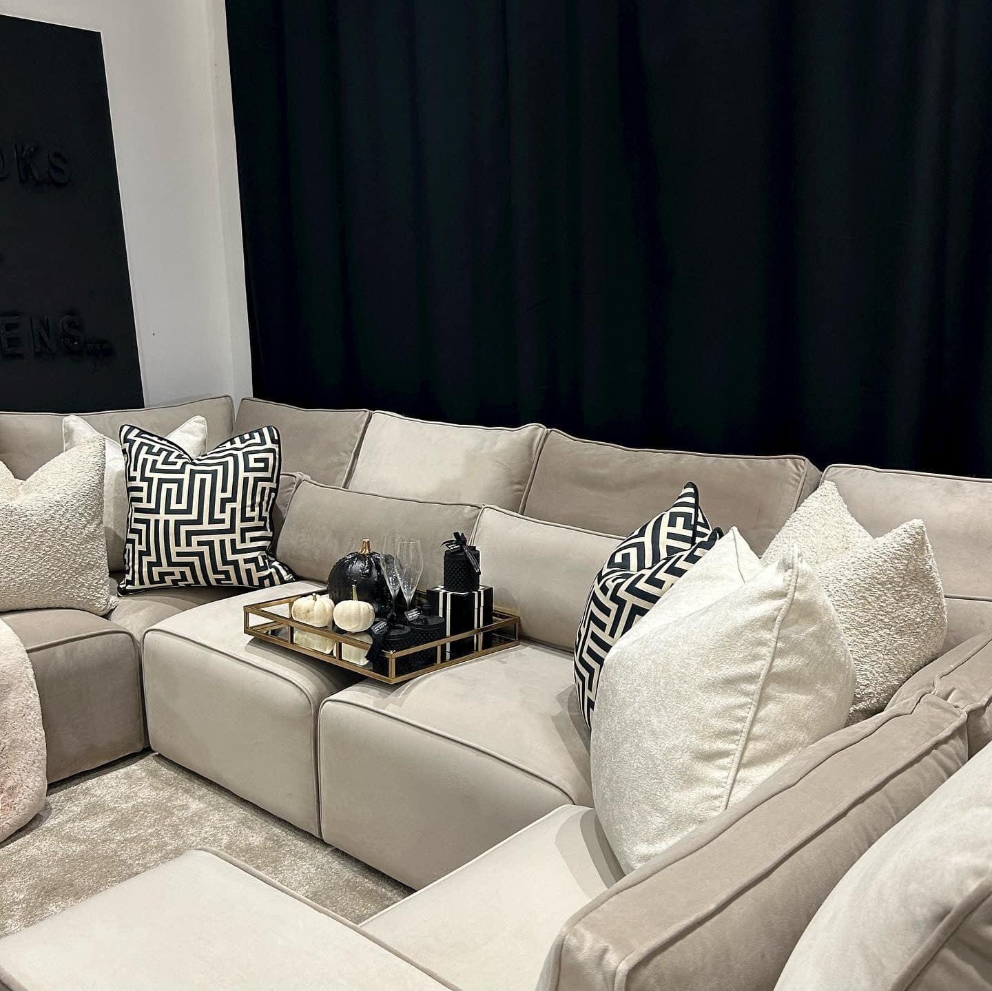 Modern U-shaped cream sofa with plush and geometric pillows, decorative tray on seat.