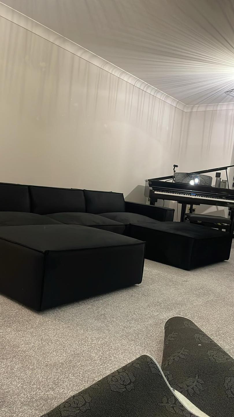 Modern black sectional sofa with ottoman on light carpet in living room, digital piano in background