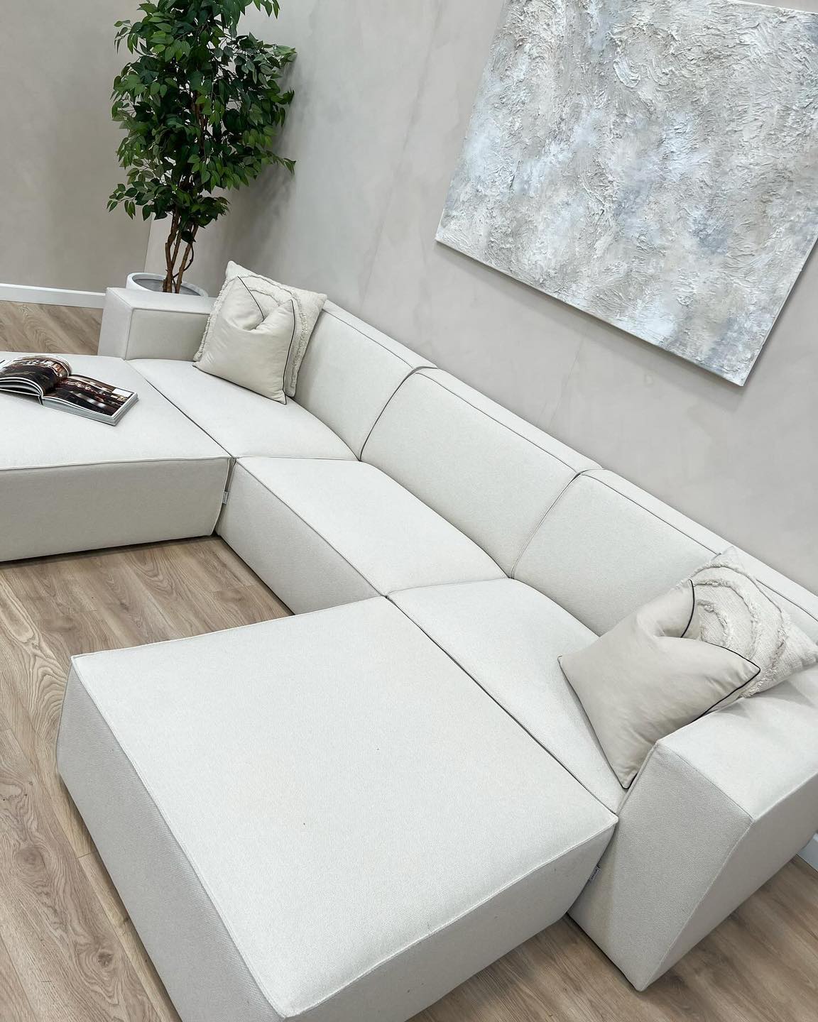 Modern ivory sectional sofa with removable footstool in a bright living room, wood floor, cushions.