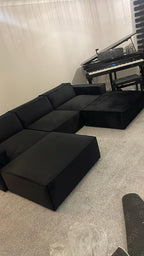 Modern black corner sofa with ottoman in a carpeted room, digital piano in background