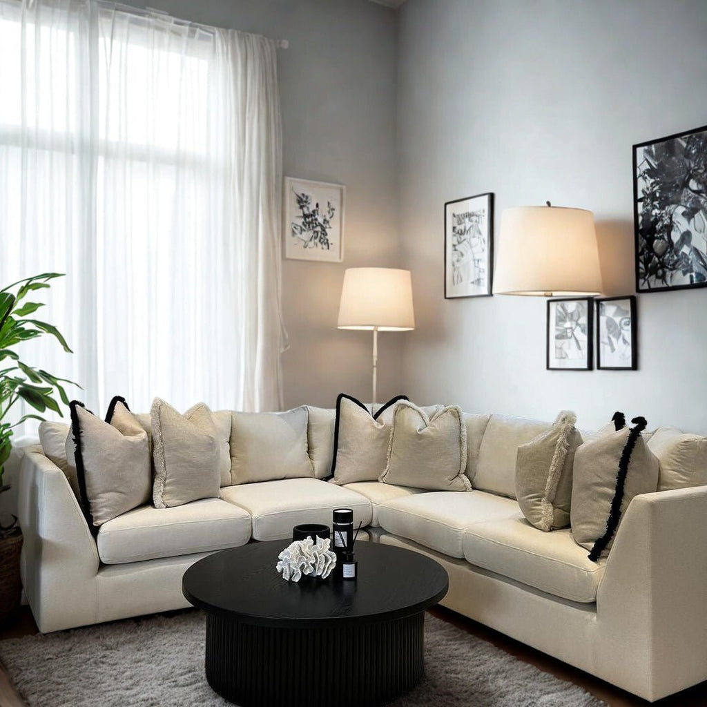 Modern living room with a cream corner sofa, plush cushions, round black coffee table, floor lamp, wall art, and large window with sheer curtains.