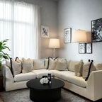 Modern living room with a cream corner sofa, plush cushions, round black coffee table, floor lamp, wall art, and large window with sheer curtains.