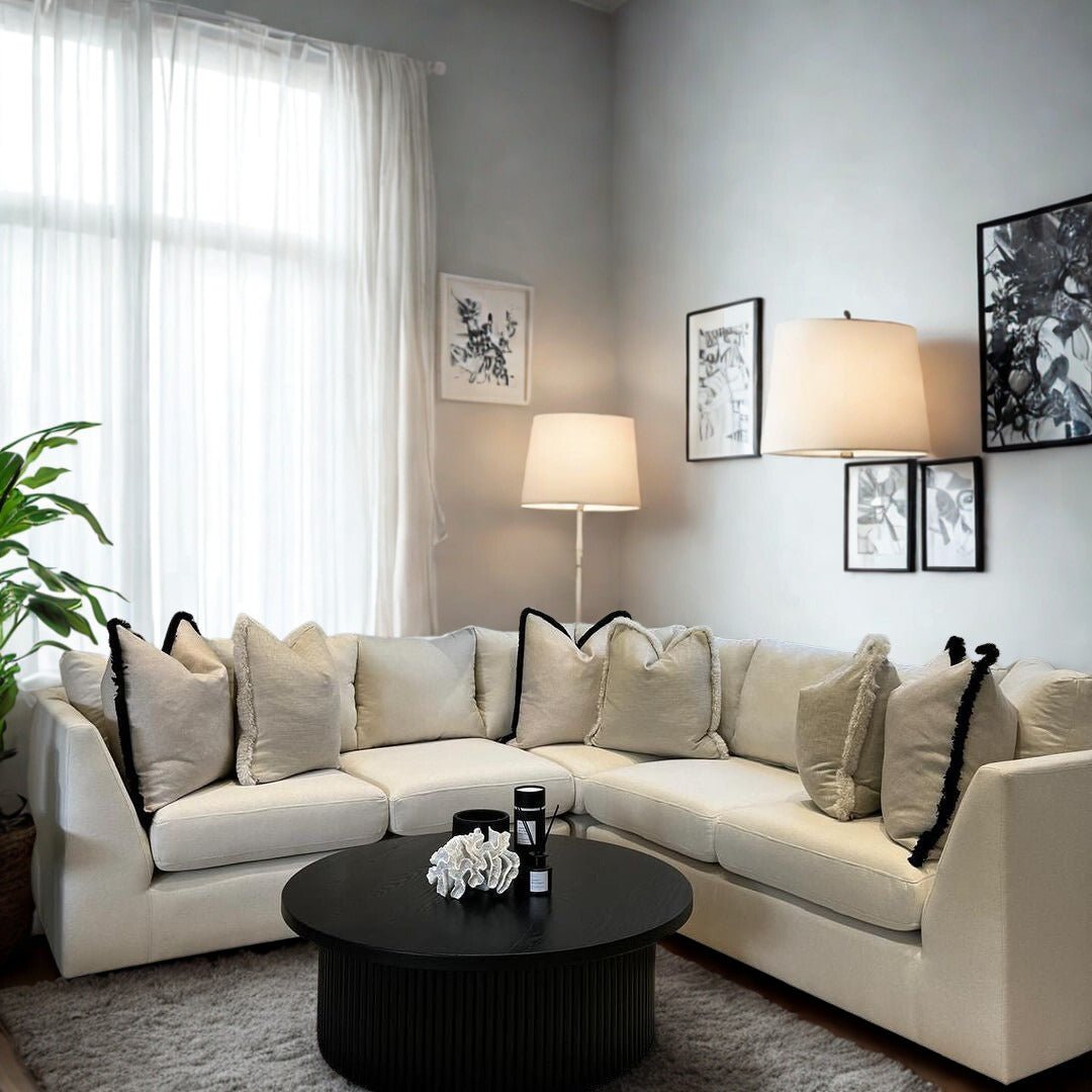 Modern living room with a cream corner sofa, plush cushions, round black coffee table, floor lamp, wall art, and large window with sheer curtains.