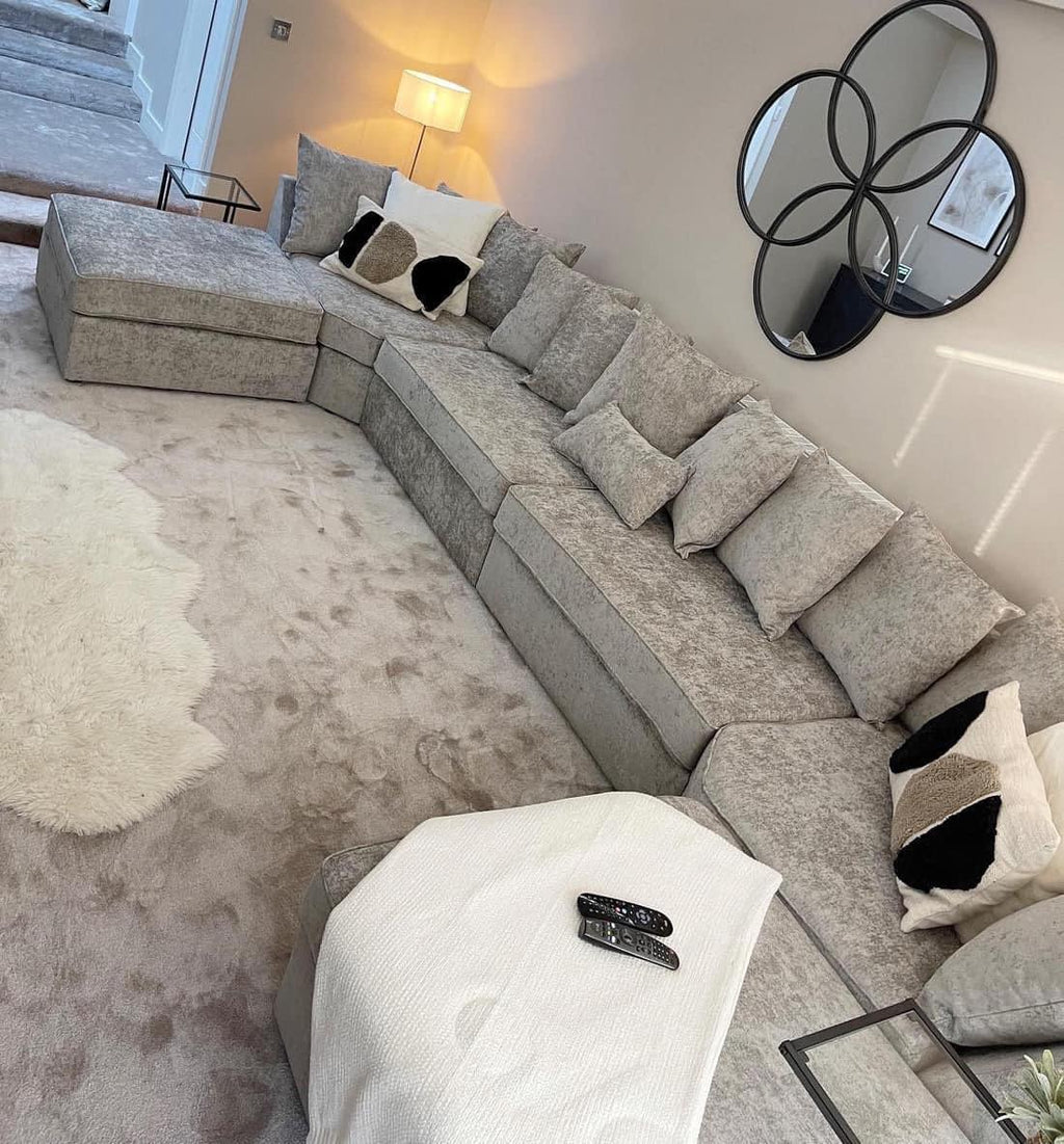 Large U-shape grey sofa with scatter cushions in a modern living room, plush carpet, and wall mirror.