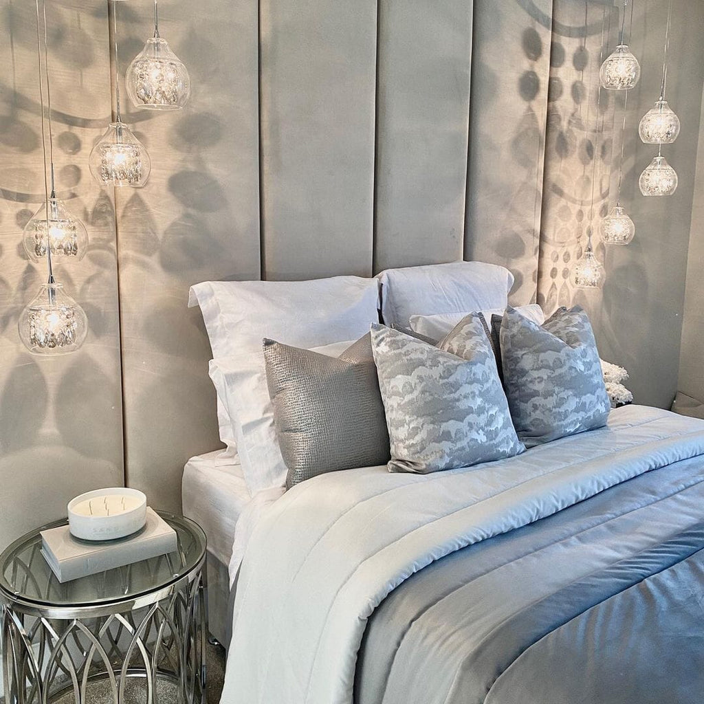 Cream velvet upholstered headboard wall panels behind a neatly made bed with decorative pillows and modern pendant lighting.