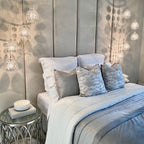 Cream velvet upholstered headboard wall panels behind a neatly made bed with decorative pillows and modern pendant lighting.