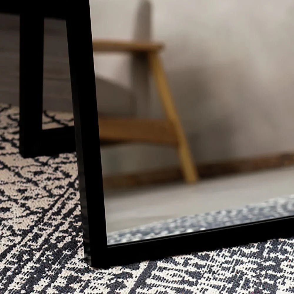 Black-framed mirror on patterned rug, reflecting wooden chair in modern living room
