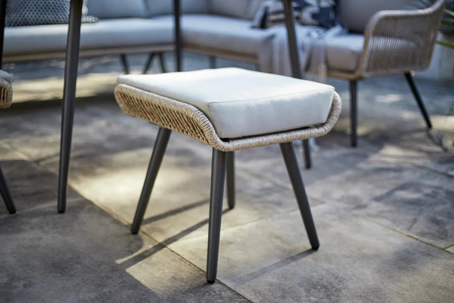 Woven outdoor stool with cream cushion and black legs on patio, matching luxury garden furniture set