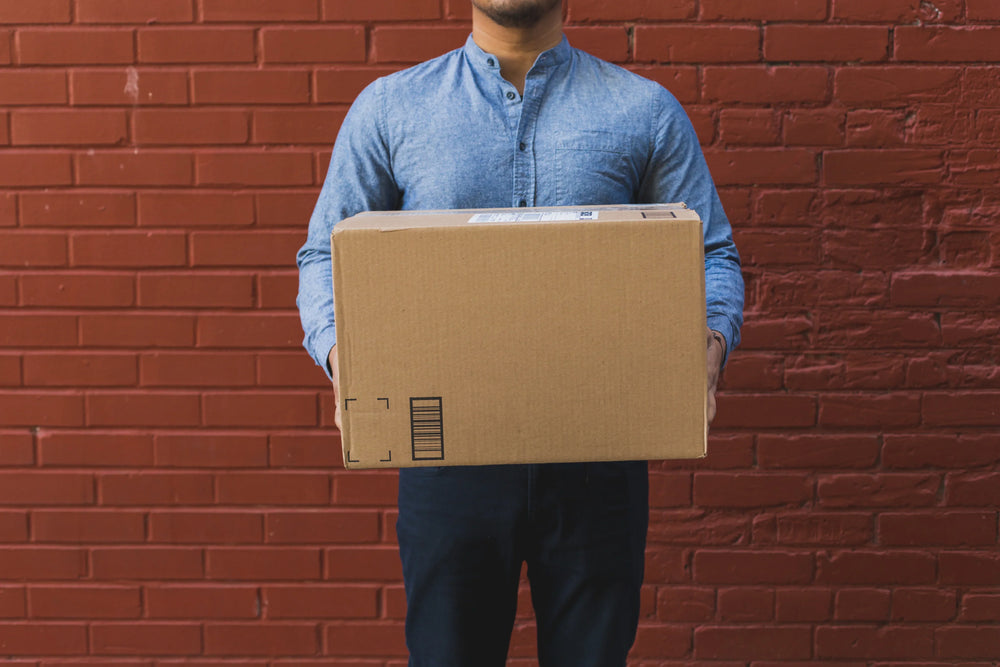 Man in blue shirt holding cardboard shipping box against red brick wall, delivery concept