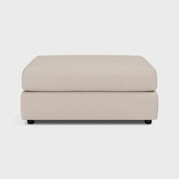 Modern beige fabric footstool ottoman with low black legs on a plain background