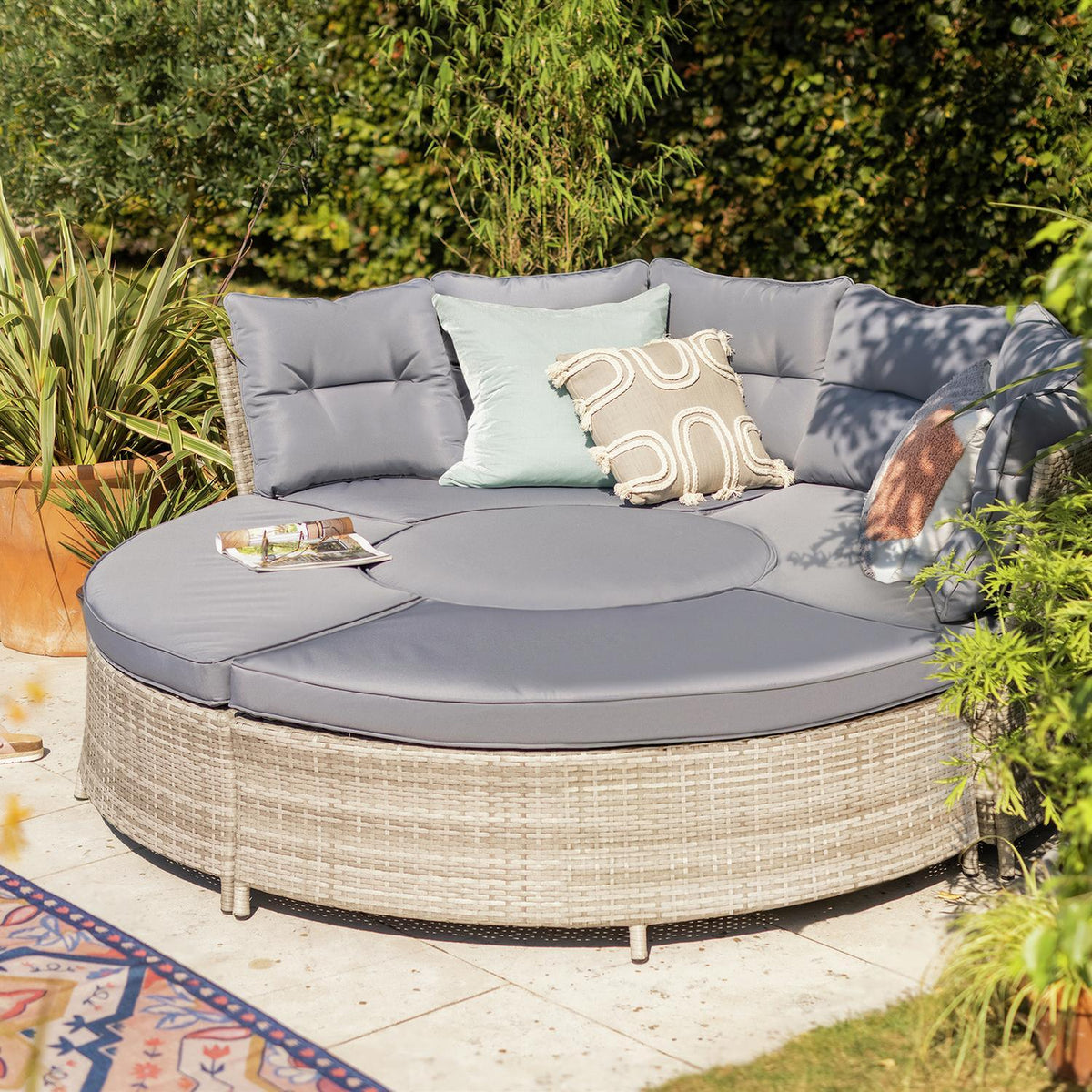 Round outdoor wicker sofa with grey cushions and decorative pillows in garden setting